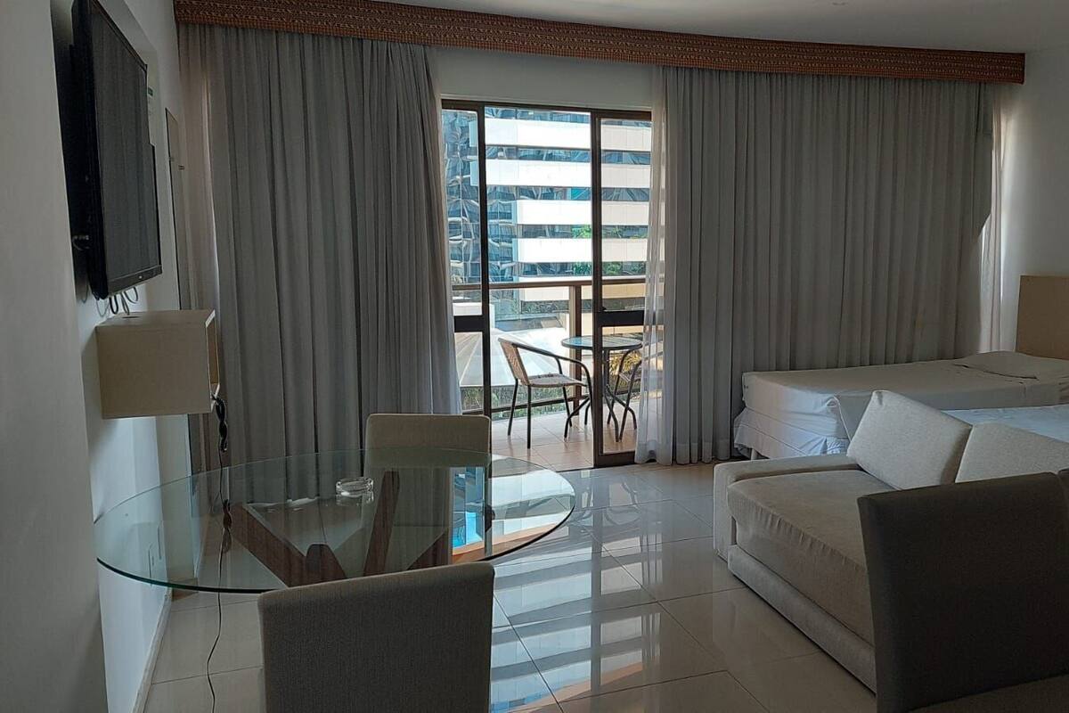 A hotel room with a glass dining table, a sofa, a bed, and a balcony with curtains.