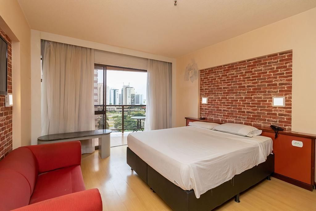 A hotel room with a large bed, a red chair, a small table, and a balcony with a city view.