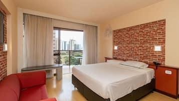 Habitación de hotel con una cama grande, una silla roja, una mesita pequeña y un balcón con vista a la ciudad.