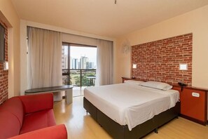 Habitación de hotel con una cama grande, una silla roja, una mesita pequeña y un balcón con vista a la ciudad.