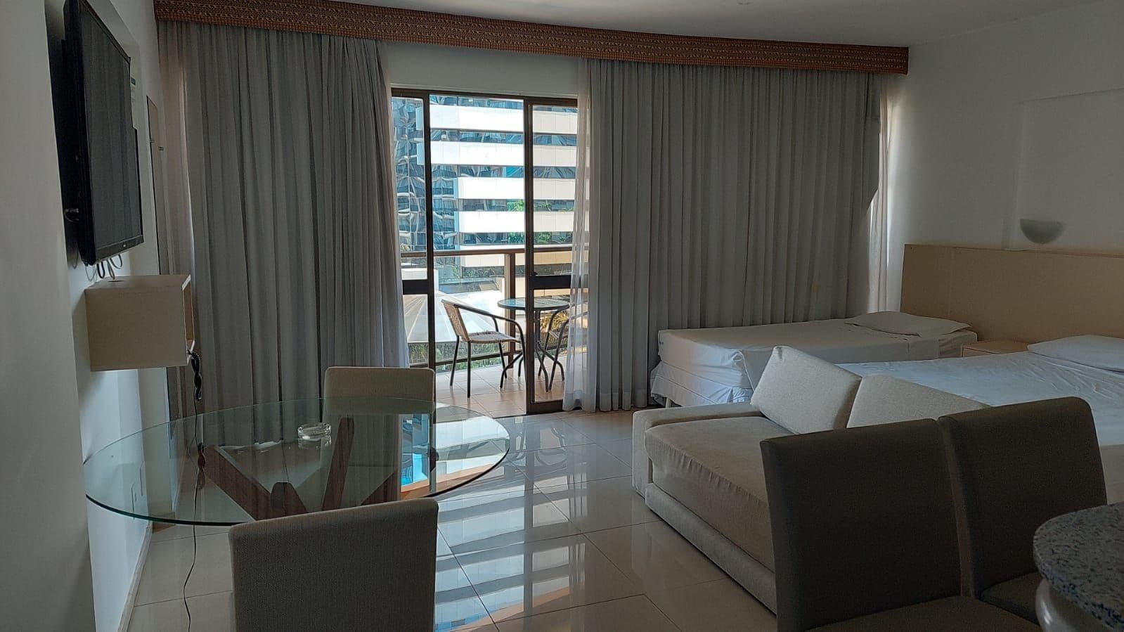 A hotel room with a glass dining table, a sofa, a bed, and a balcony with curtains.
