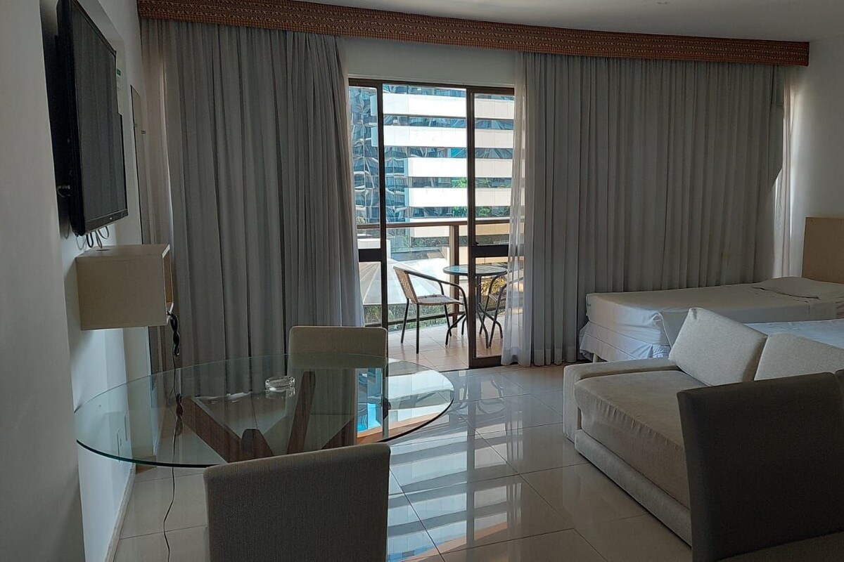 Una habitación de hotel con una mesa de comedor de cristal, un sofá, una cama y un balcón con cortinas.