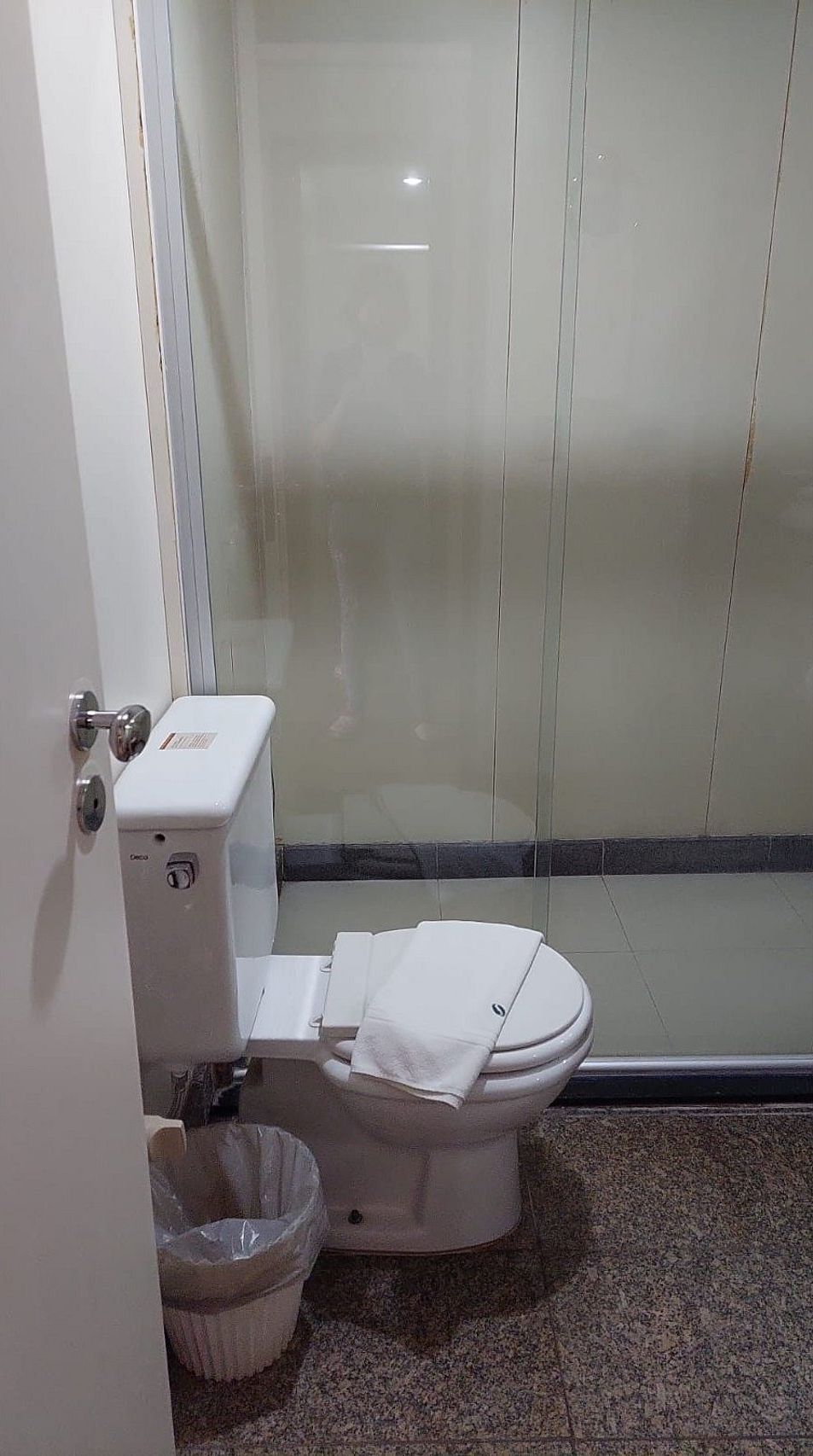 A small, enclosed bathroom with a toilet, a trash bin, and a glass shower enclosure.