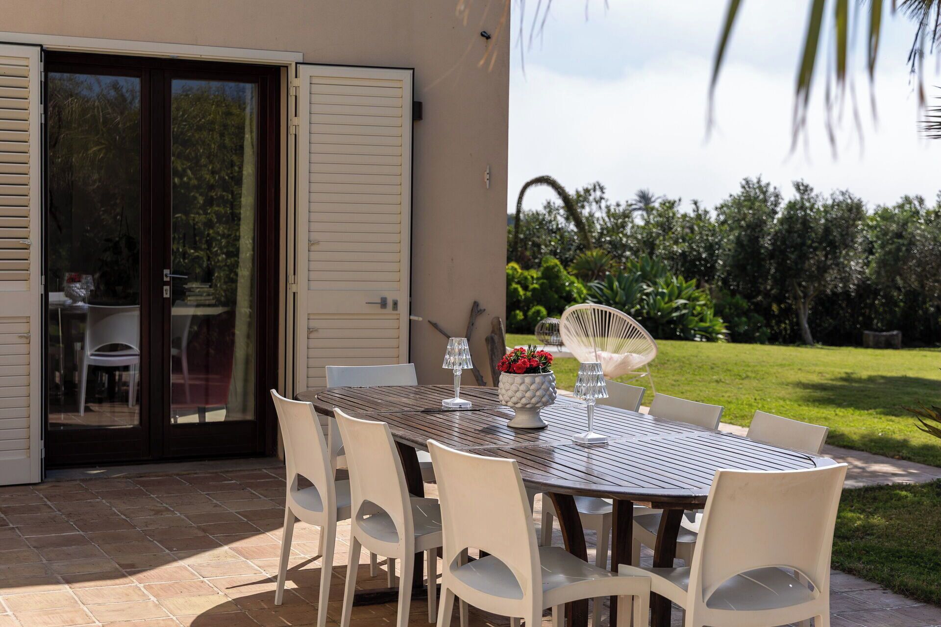 An outdoor dining area with a table and chairs, a glass door, and a view of a garden.