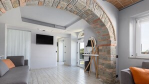 Un salón espacioso con un arco de piedra, sofás grises con cojines naranja, un televisor de pantalla plana montado en la pared y un caballete de madera apoyado en la pared.