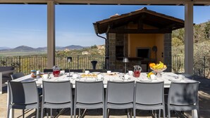 Un área de comedor al aire libre con una mesa dispuesta para seis personas, rodeada de sillas, con vistas a un paisaje de colinas y un edificio.