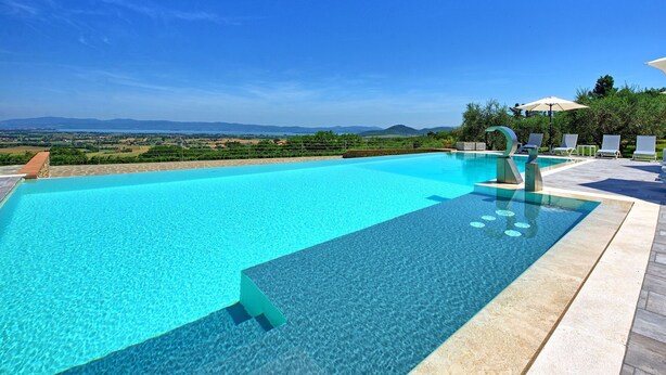 Una piscina infinita de color azul claro con vistas a montañas distantes, rodeada de sillas de descanso y una sombrilla blanca.