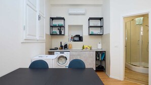 A compact kitchen with a washing machine, a sink, and a microwave. There are two chairs at a table, and a glass shower door is visible. - Dominium Premium Location Suites Studio apartment (Dubrovnik)
