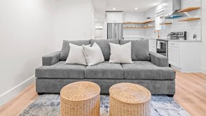 Una sala moderna con un sofá gris, dos mesitas de mimbre y una cocina al fondo.
