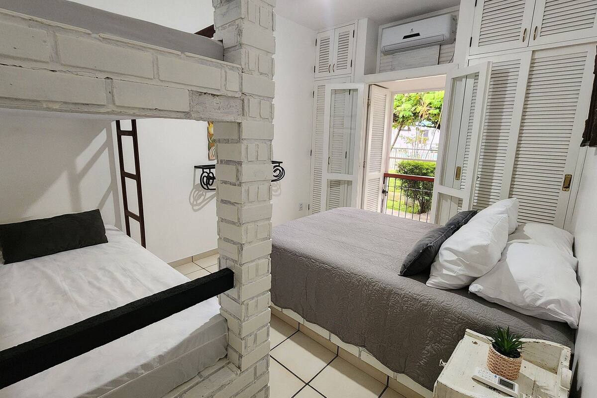 A room with a bunk bed, a single bed, a small table with a plant, and a window with shutters.