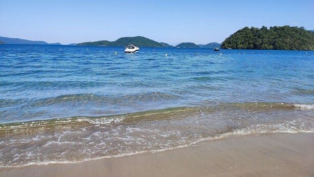 A beach with clear water, a boat, and distant hills. - Angra Temporada (Angra dos Reis)