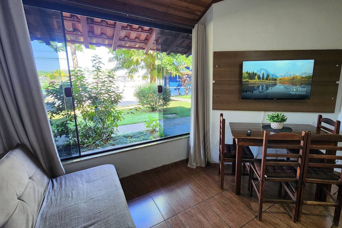 A room with a large window, a TV mounted on the wall, a dining table with chairs, and a sofa.