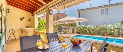 Un área de comedor al aire libre techada con vista a la piscina, una mesa con alimentos para el desayuno y un muro de piedra.