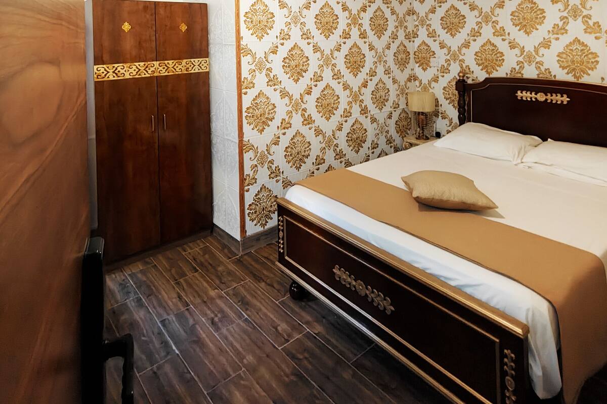 A bedroom with a wooden wardrobe, a bed with a beige bedspread, and patterned wallpaper.