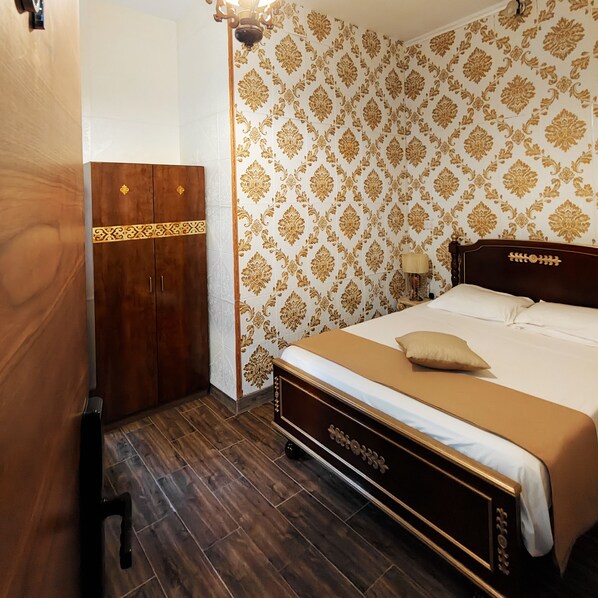 Un dormitorio con un armario de madera, una cama con colcha color crema y papel tapiz estampado.