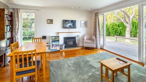 Amplia sala de estar con una mesa de comedor de madera, sillas, chimenea, estantería y ventanas grandes con vistas al exterior.