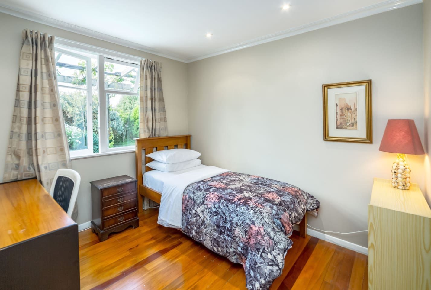 A bedroom with a wooden bed, a desk, a chair, a dresser, a window with curtains, a bedside lamp, and a framed picture on the wall.