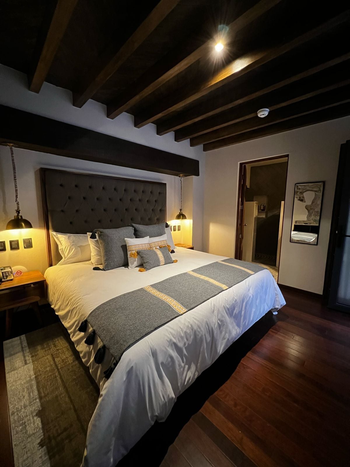 Une chambre à coucher avec un grand lit, des poutres en bois, une tête de lit, des lampes de chevet, une table de chevet et une porte donnant sur une autre pièce.