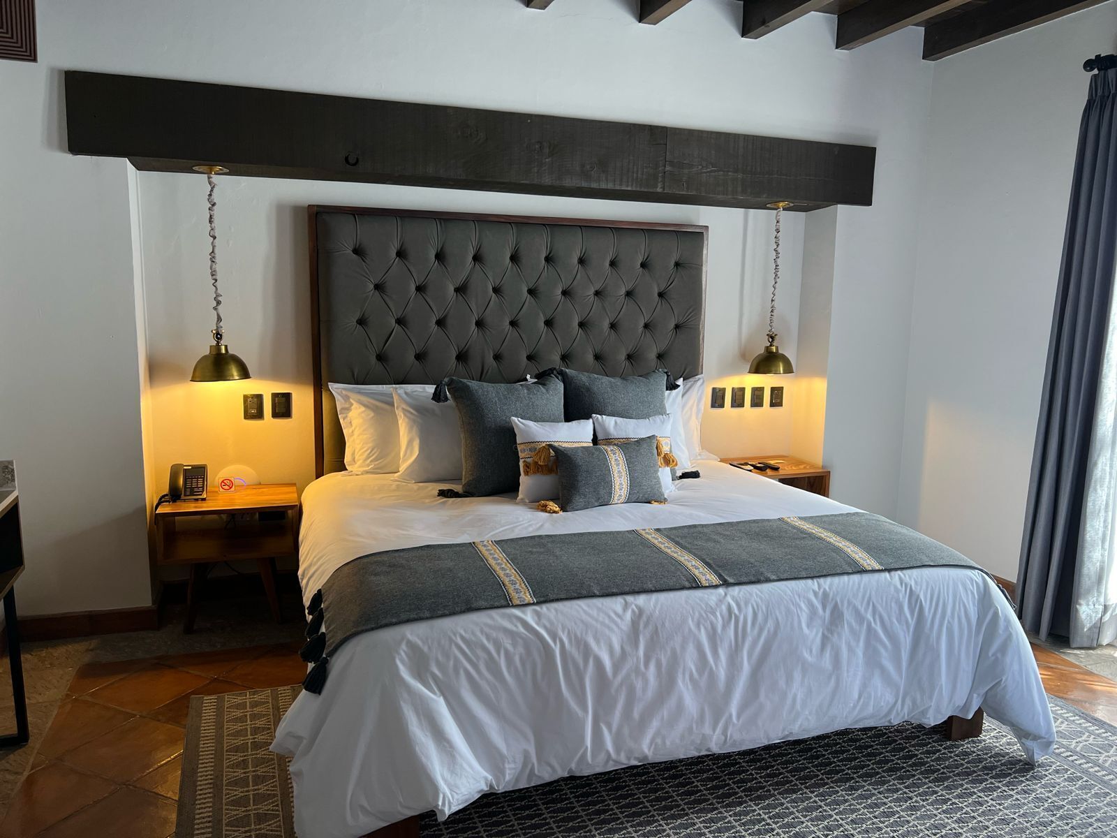 A bedroom with a tufted headboard, a bed with white and grey bedding, two bedside tables with lamps, and a patterned rug.