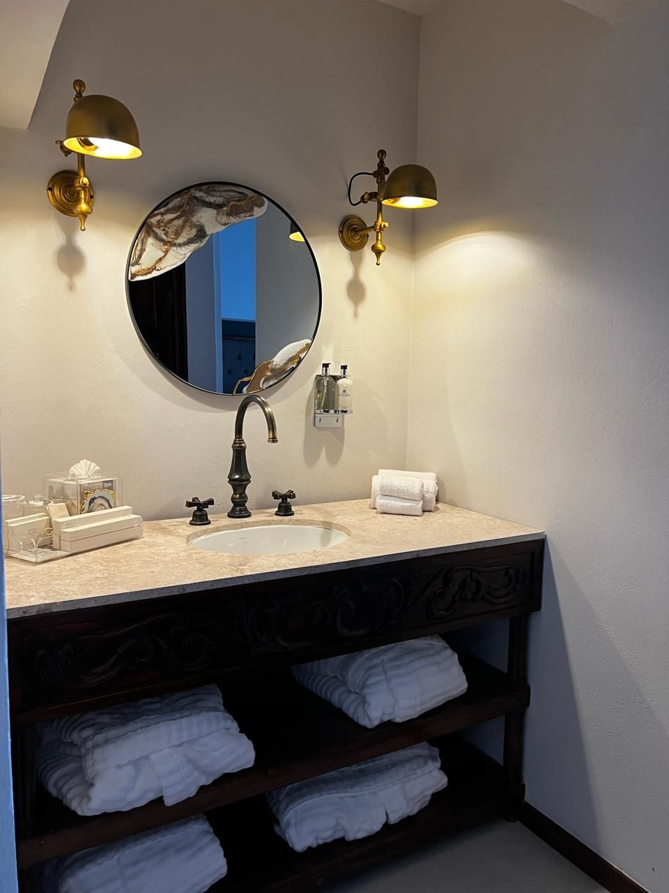 Une salle de bain avec un miroir rond, deux lampes fixées au mur, un lavabo et un porte-serviettes avec des serviettes pliées.