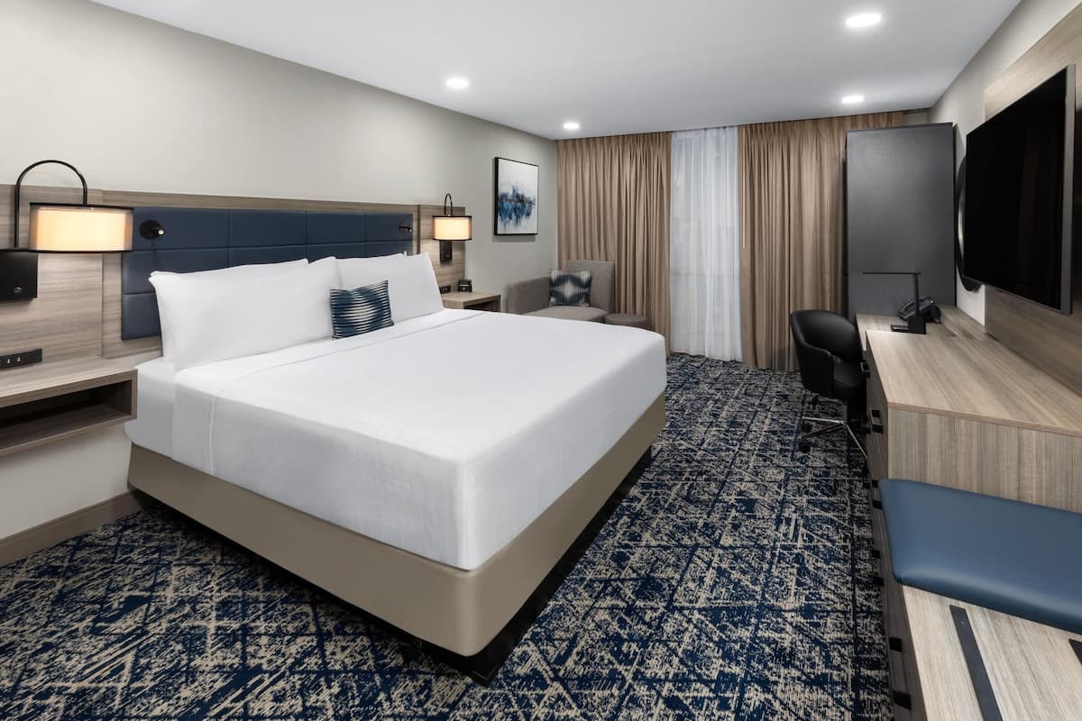 A hotel room with a large bed, a desk with a chair, a flat-screen TV, and a wall-mounted TV.