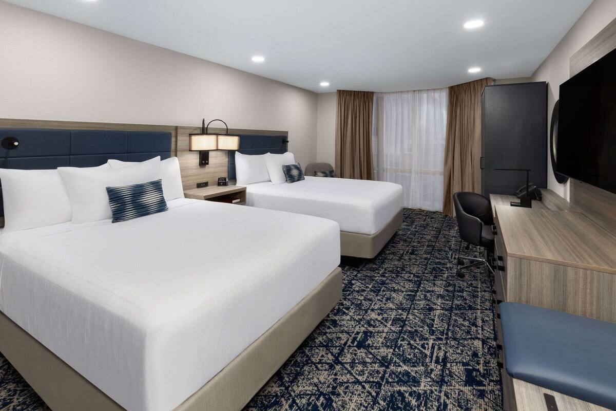 A hotel room with two beds, a desk, a chair, a TV, and a large window with curtains.