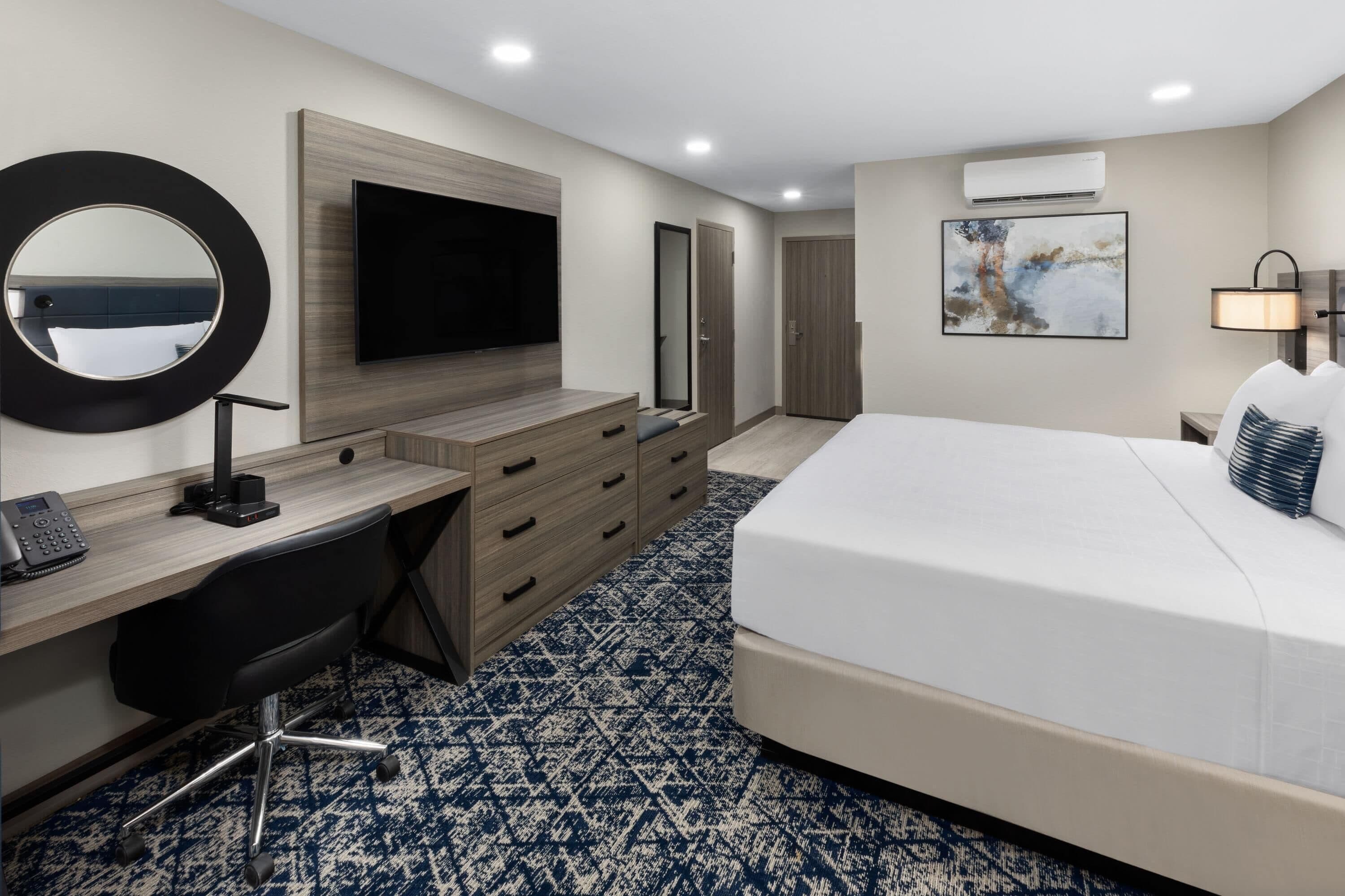 Habitación de hotel con cama, escritorio con computadora, televisor, espejo, armario y unidad de aire acondicionado.