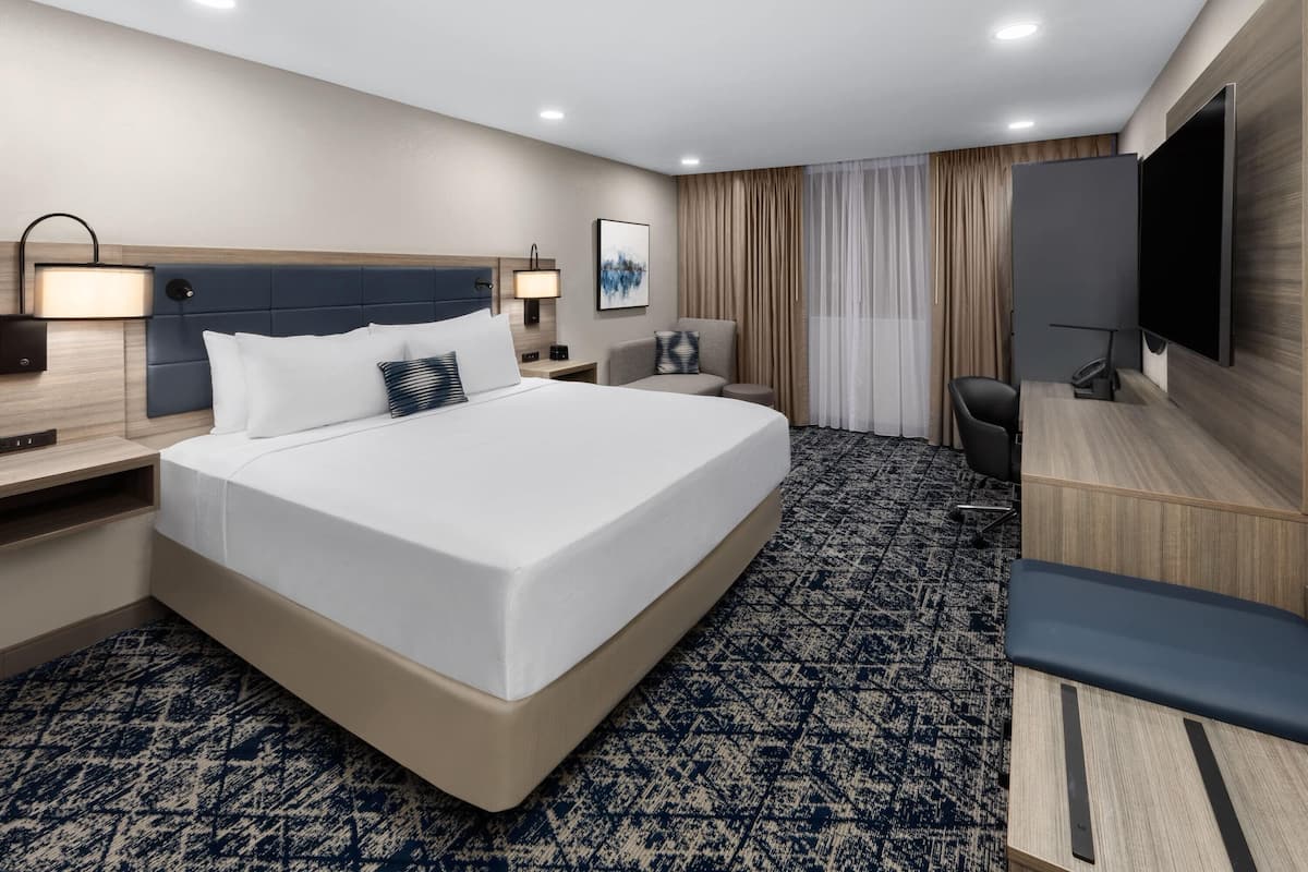 A hotel room with a large bed, a desk, a chair, a TV, and a window with curtains.