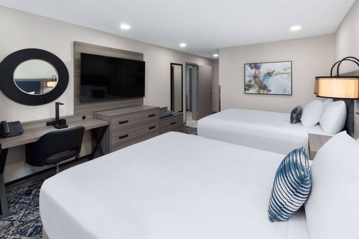 A hotel room with two beds, a desk with a chair, a desk lamp, a telephone, a large mirror, and a painting on the wall.