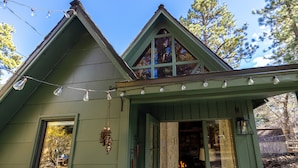 Une maison verte avec un toit triangulaire, un porche à l’avant et une fenêtre ornée d’une décoration suspendue.