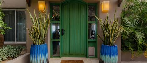 Una puerta verde con un ventanal pequeño, flanqueada por dos macetas azules grandes con plantas altas.