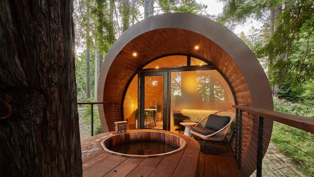 Une cabane circulaire en bois, dotée d’un jacuzzi et d’un espace salon sur une terrasse, le tout entouré d’arbres.
