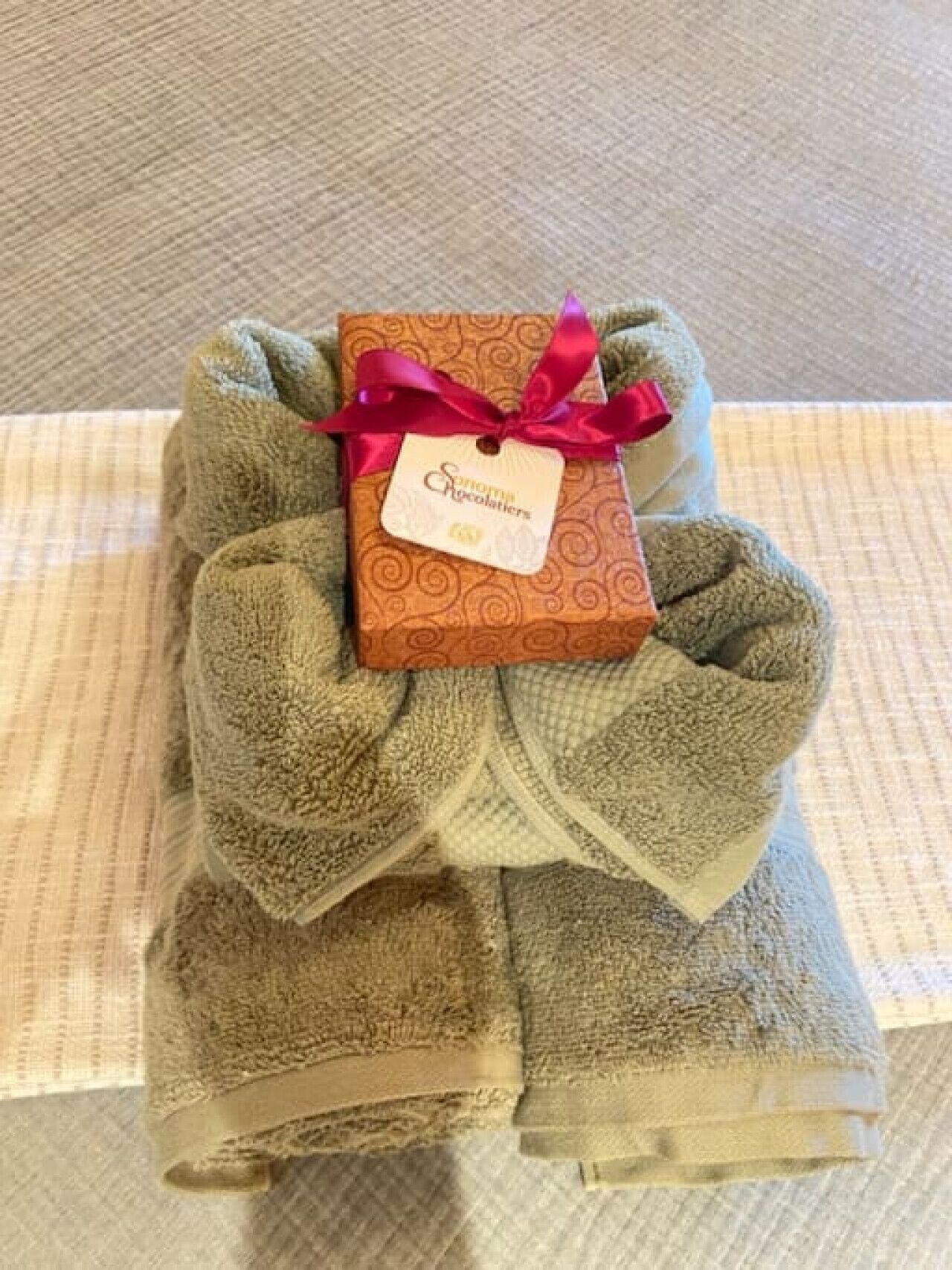 A neatly folded towel bundle with a gift box on top, tied with a ribbon, placed on a patterned surface.