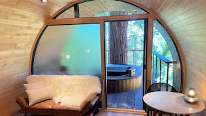 Une pièce chaleureuse avec un plafond en bois aux lignes courbes, un lit recouvert d’une couverture blanche et moelleuse, une petite table ronde agrémentée d’un objet décoratif, et une vue sur un balcon doté d’une terrasse en bois et d’un arbre.