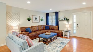 A living room with a brown leather sofa, a blue armchair, and a wooden coffee table. There is a white door, a blue curtain, and a small white table with a lamp. - Virginia Escape w/ Patio & Fire Pit, Near Hiking! (Wytheville)