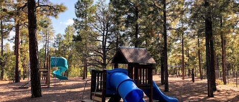 A playground with a blue slide and a wooden playhouse in a wooded area.