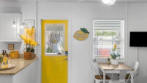 Una porta gialla brillante con un oblò in vetro satinato, una parete bianca con il cartello 'SQUEEZE THE DAY', un tavolo di legno con sedie e una ventola a soffitto.