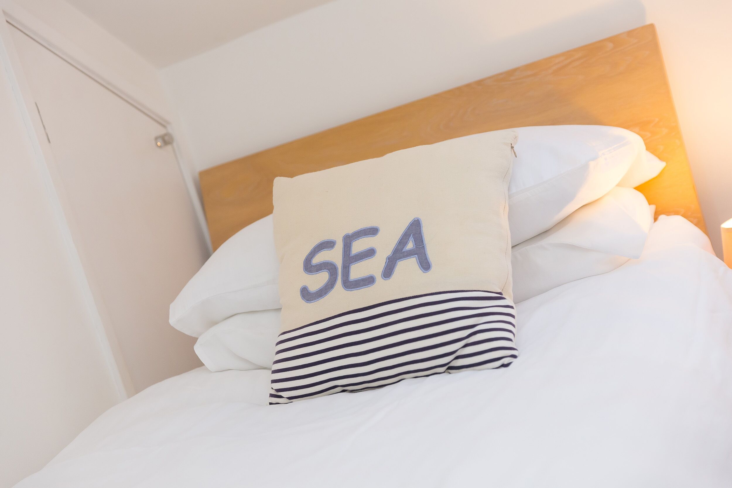 Een net opgemaakt bed met witte beddengoed en een kussen waarop de tekst 'SEA' staat.
