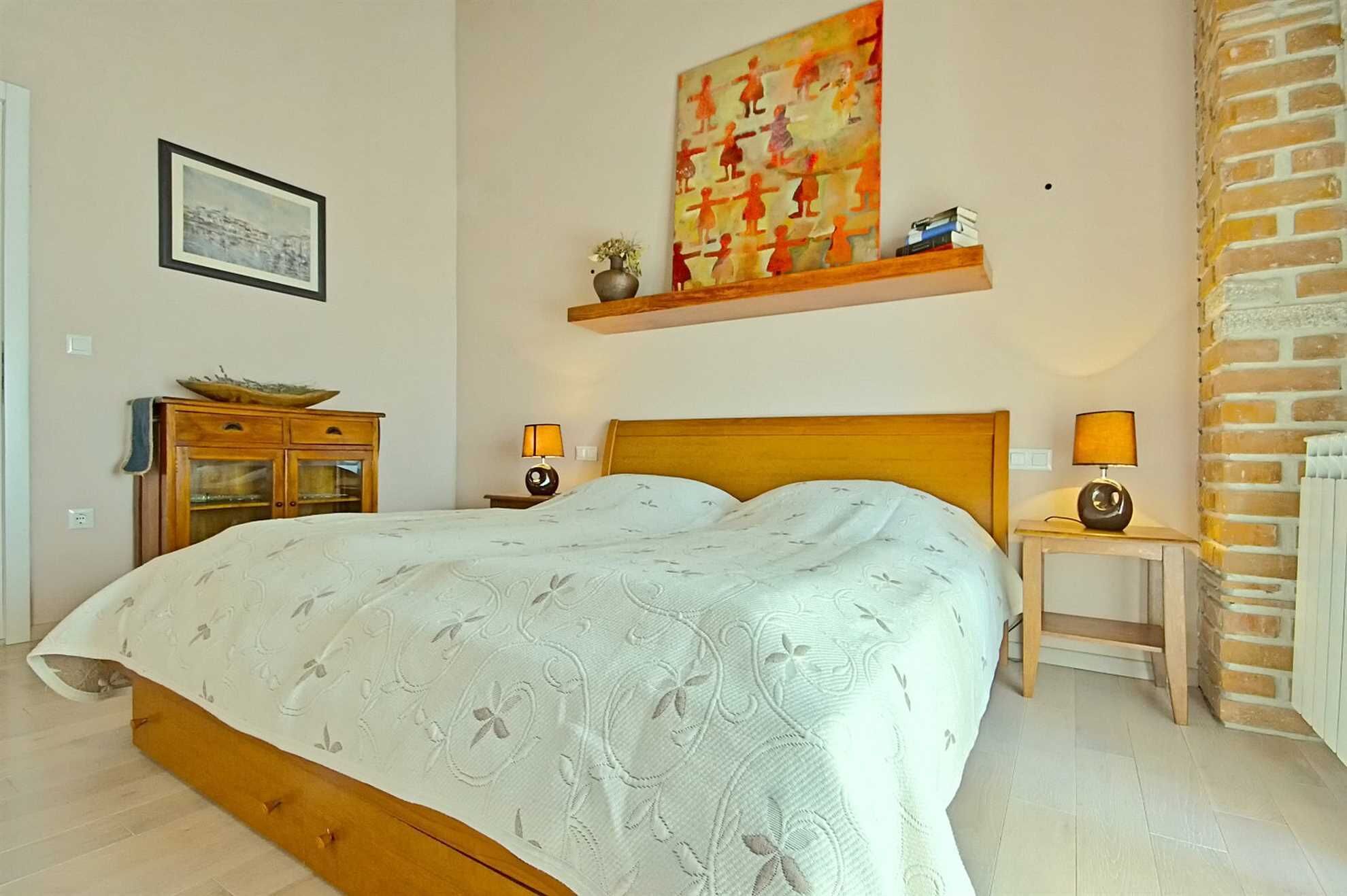 Une chambre avec un lit en bois, deux tables de chevet avec des lampes, une étagère murale avec des livres, un tableau encadré et un mur en briques.