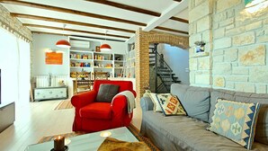 Una sala de estar con un sofá rojo, uno gris y una mesa de centro de vidrio. Se observa una estantería y una escalera al fondo.