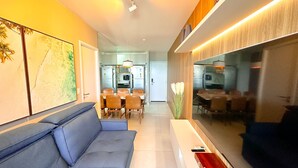 A modern living room with a blue sofa, a dining area with a table and chairs, and a kitchen visible through a glass partition. - Edifício Time (Maceió)