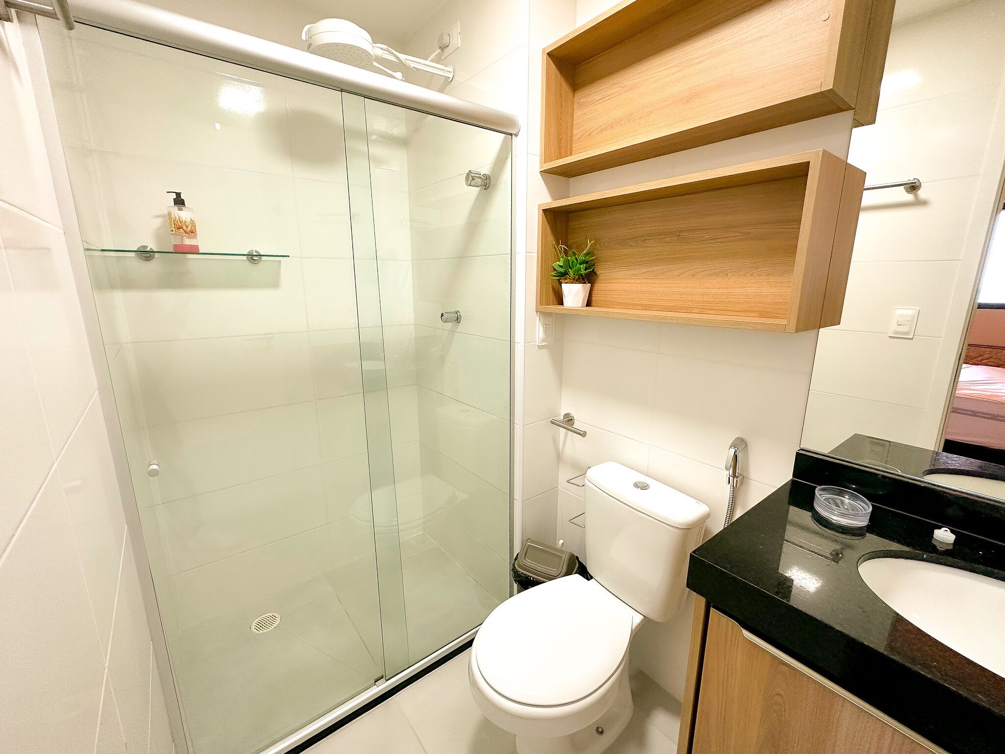 Un baño moderno con ducha envuelta en vidrio, inodoro blanco, lavabo negro y estantes de madera sobre el inodoro.