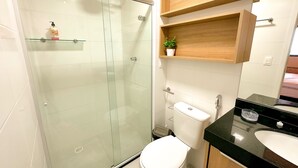 A modern bathroom with a glass-enclosed shower, a white toilet, a black sink, and wooden shelves above the toilet. - Edifício Time (Maceió)