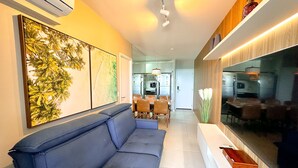 A modern living room with a blue sofa, a dining area with chairs, and a kitchen visible through an open door. - Edifício Time (Maceió)