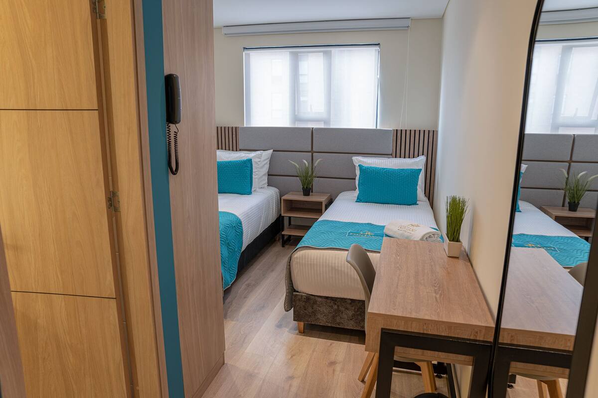 A compact hotel room with two beds, a desk, and a small table.