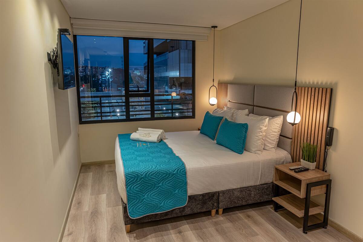 A modern bedroom with a large window, a bed with blue and white bedding, a bedside table with a plant, and a nightstand with a phone.