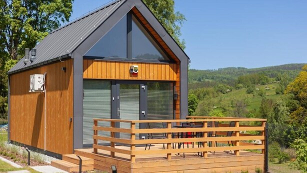 A modern cabin with a wooden deck and a large window, set against a scenic hillside backdrop.