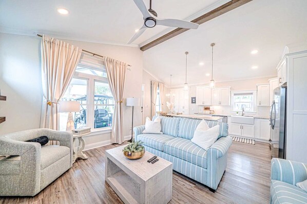 A spacious living room with a sofa, armchair, and coffee table. A kitchen with white cabinets and a refrigerator is visible in the background. - 232 Barracuda Dr. - 3 Bedrooms - Includes 2 Golf Carts & 2 Sets of Beach Chairs (Panama City Beach)