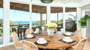 Une table ronde en bois, dressée pour le repas, entourée de chaises en osier, un vase de fleurs jaunes et une vue sur la mer par de grandes fenêtres.