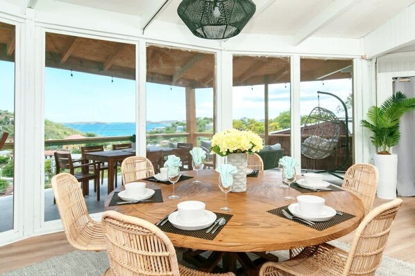 A round wooden table set for dining with wicker chairs, a vase of yellow flowers, and a view of the sea through large windows.